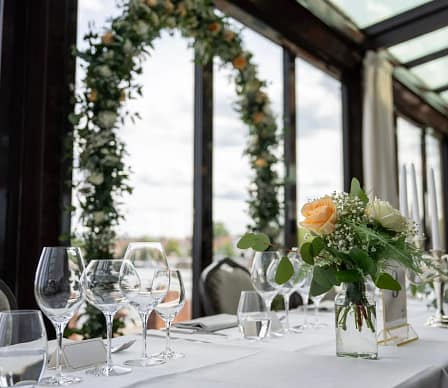 Festdukning på Slottsholmen med vita linnedukar, blomsterarrangemang, vinglas och blomsterbåge med utsikt över vattnet.